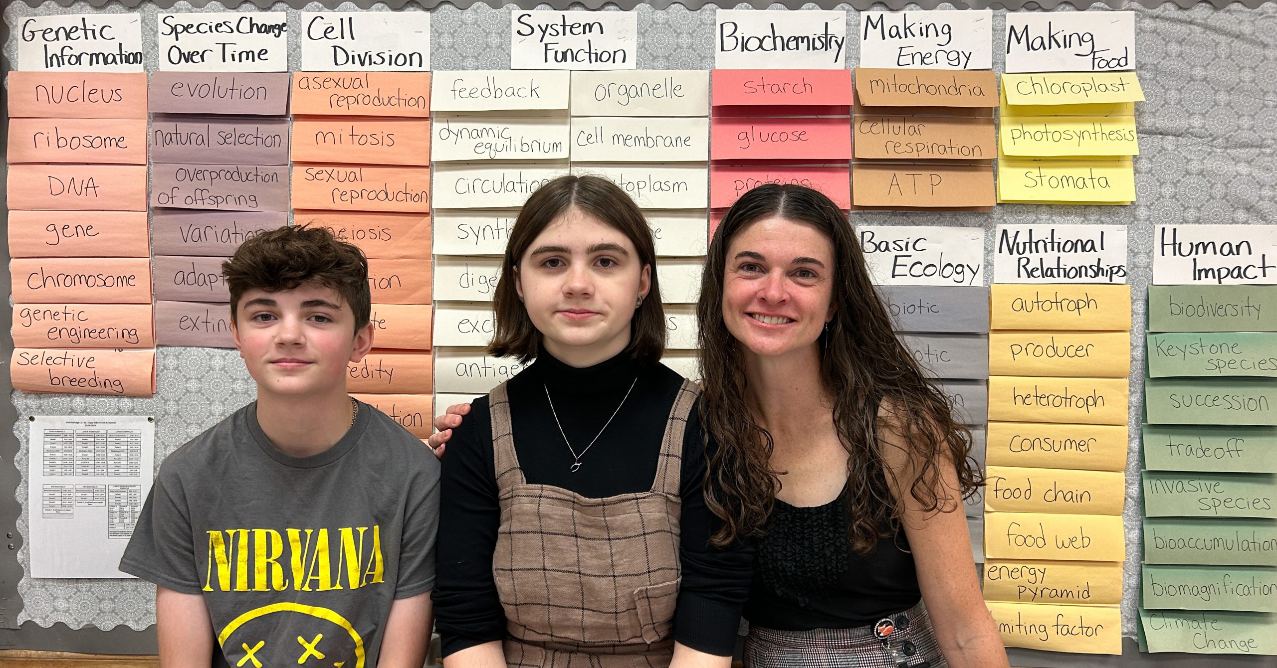 Teacher and two students in front of biology vocabulary bulletin board.