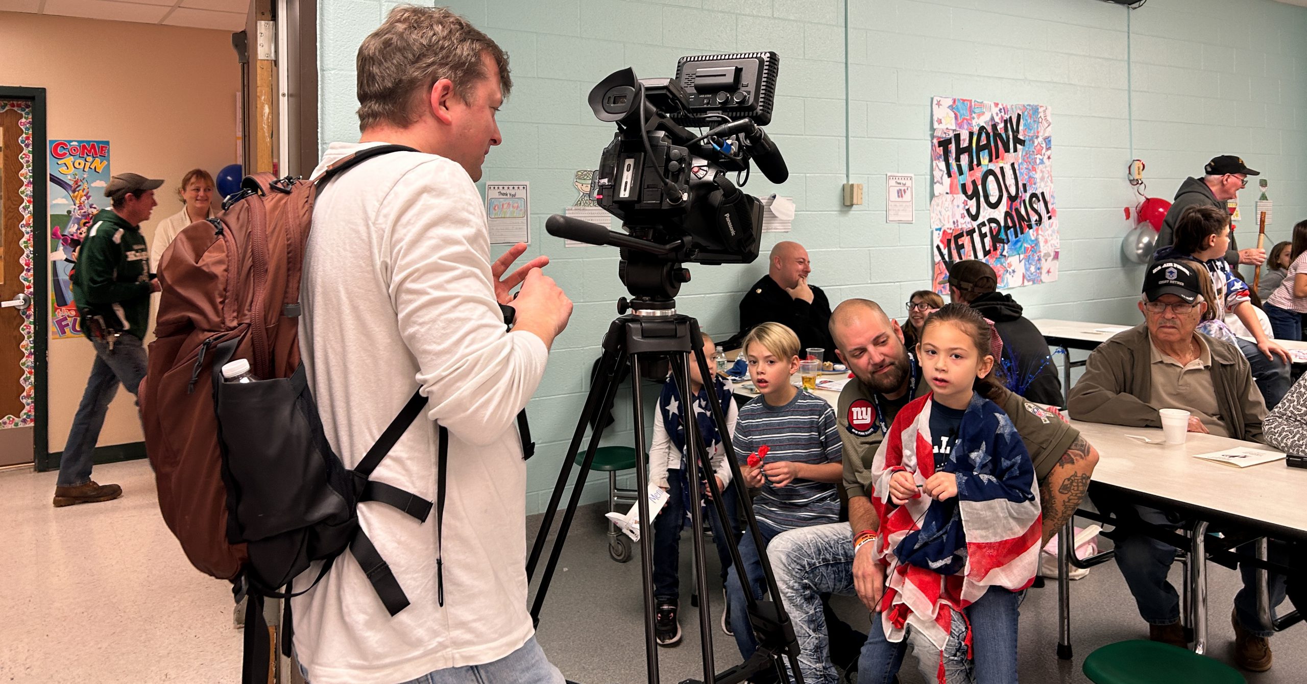 Videographer stands behind camer. Children and veterans are seated at table.