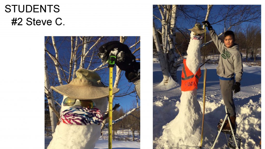 A very tall and long snowman that's all in one piece, wearing a hat and sunglasses and a colorful mask over its mouth and orange civil air patrol vest. Student who made snowman is holding a tape measure near it and standing on a step stool to reach top. It measures a few inches taller than 7 feet.