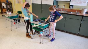 two students standing at desk with their hands on a tablet creating code