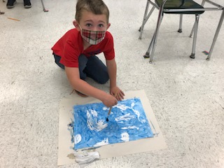 1st grade student in a red shirt and red and white mask looks up from the floor, where he's swirling a paint brush to make clouds on paper