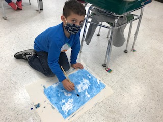1st grade student in a blue shirt and black mask looks up from the floor, where he's painting a sky on paper