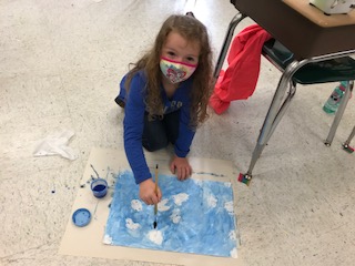 1st grade student in a blue shirt and white mask looks up from the floor, where she's painting clouds on paper