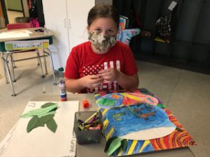 student wearing a mask sits in front of her goldfish drawing and is about to paste leaves on it