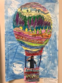 drawing of a hot air baloon with yellows, greens, purples, pinks. The baloon has a student picture in the basket and is up above most of the clouds