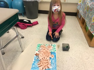 kindergarten student sits on floor in front of her painting of a teddy bear