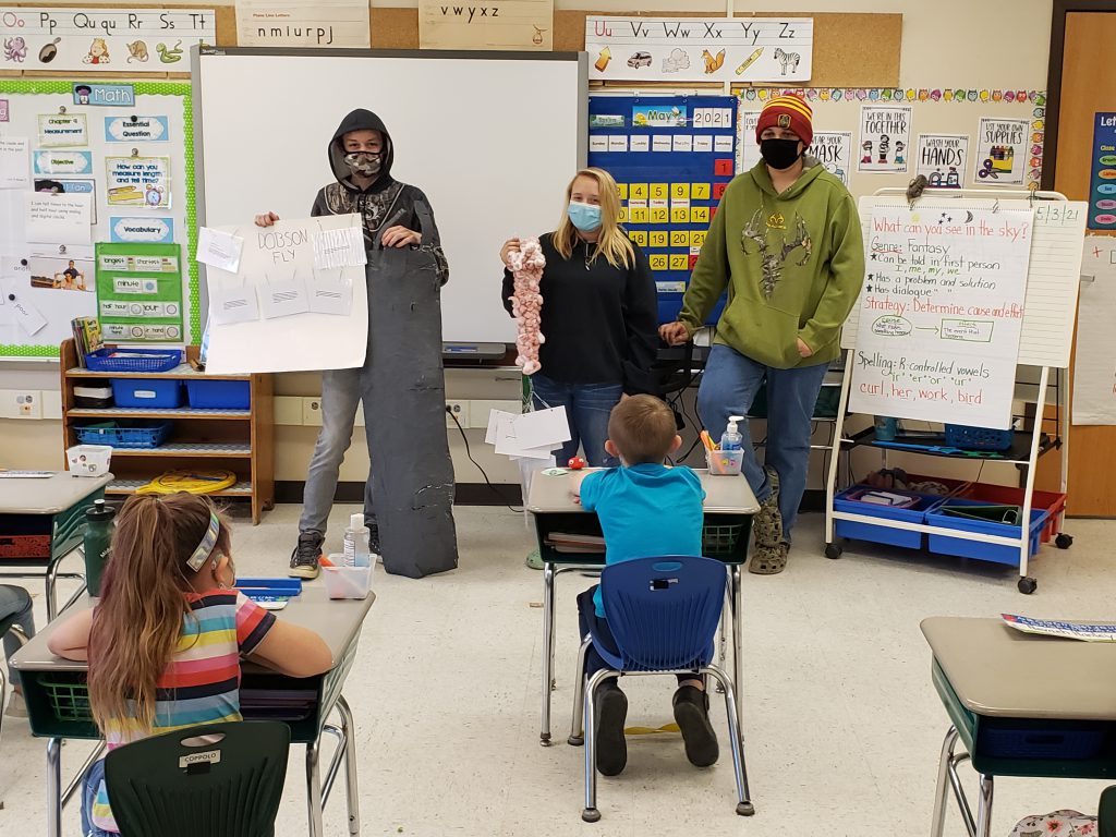 three students at front of class holding up their projects for younger students to see