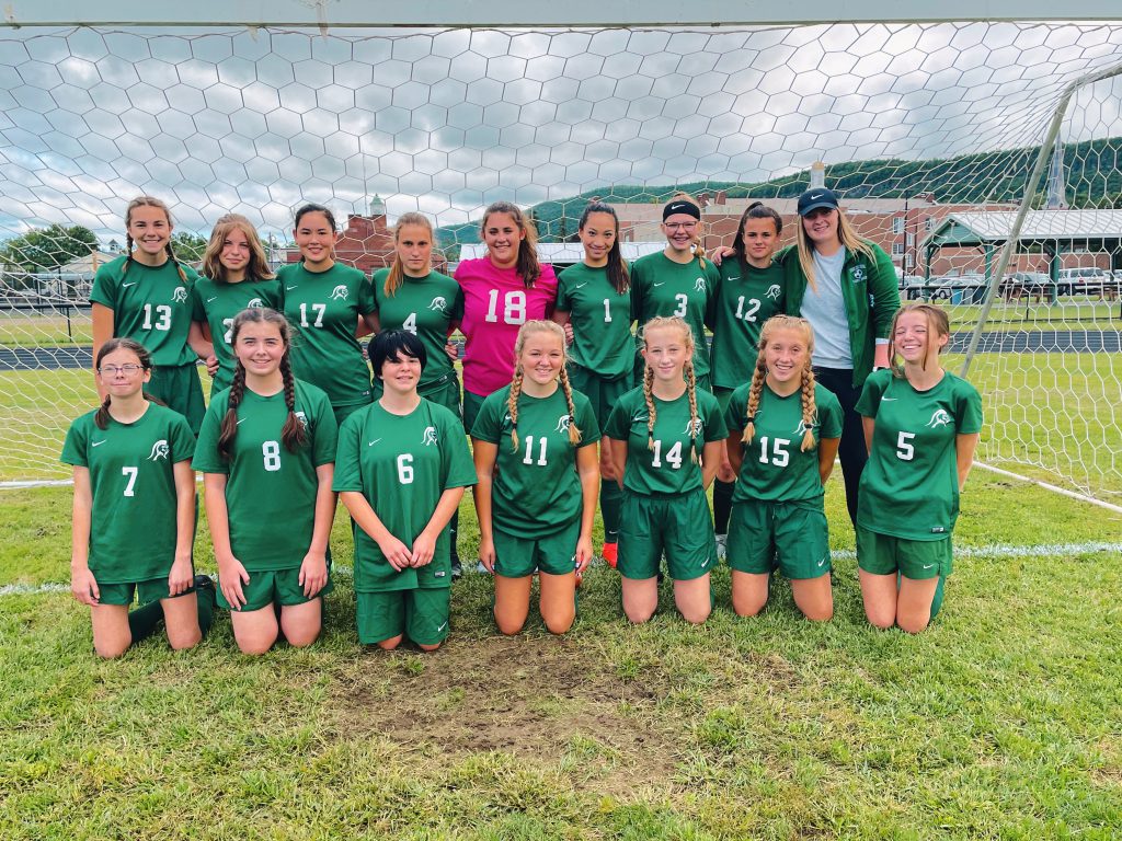 15 girls soccer players and one coach standing inside of the goal some are standing and others are kneeling in front