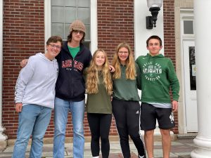 Five sophomores are standing up leaning into each other with their arms around each other outside on the steps of the high school