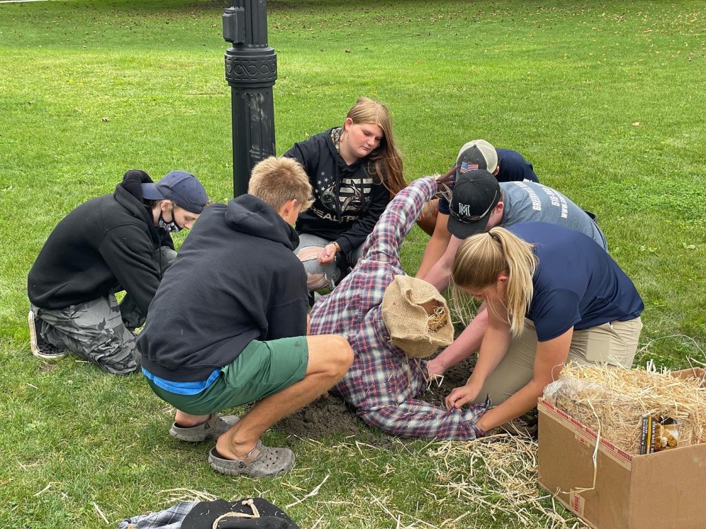 five students and a teacher are surrounding their scarecrow and stuffing it with hay