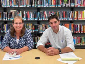 two adults sitting at a table with their hands clasped in front of a bookshelf