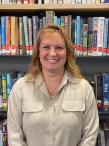 woman with feathered hair smiling in front of a book case, wearing a cream button down shirt