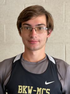 student wearing glasses and a cross country uniform standing in front of a cream colored wall