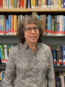 woman with curly short hair and dark rimmed glasses standing in front of a book case wearing a black and white print shirt