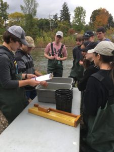 students standing near the Schoharie Creek at a table with buckets, one student is holding a clip board and the other students are looking at him