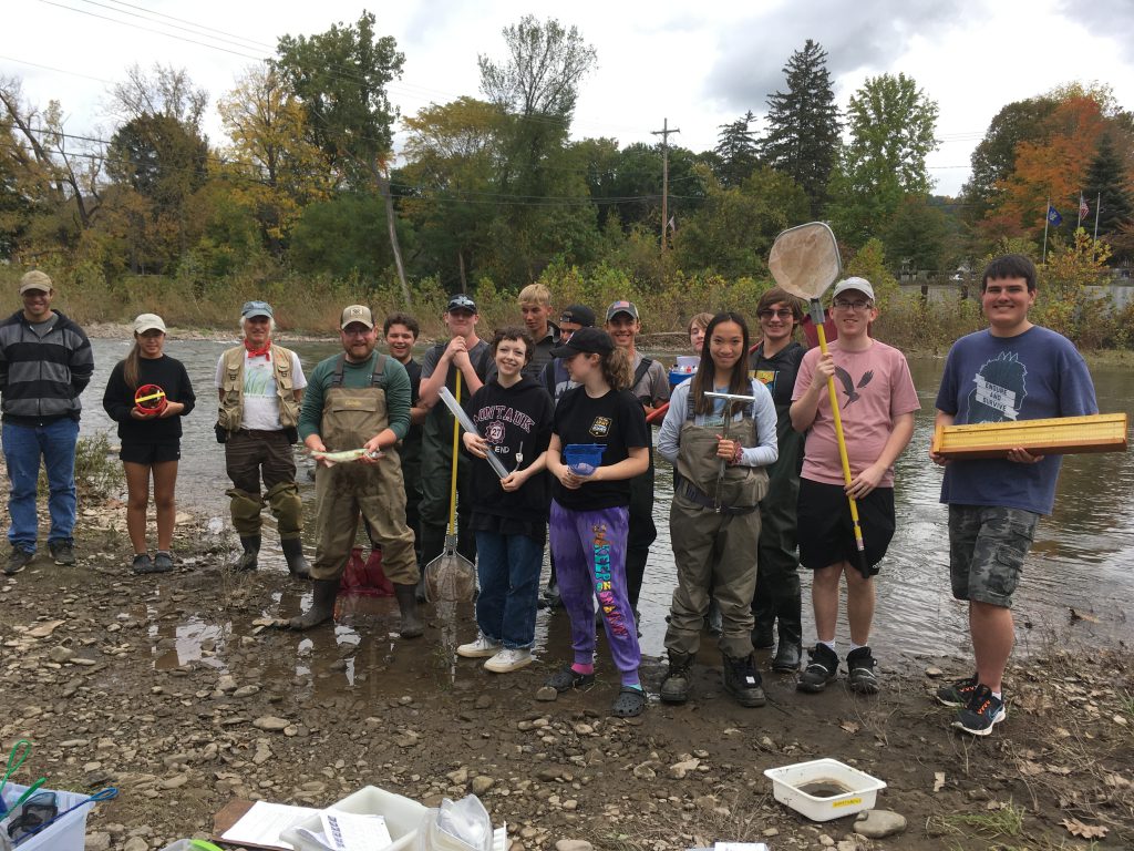 14 students and two environmental specialists stand near the Schoharie Creek holding up equipment used in a water quality study