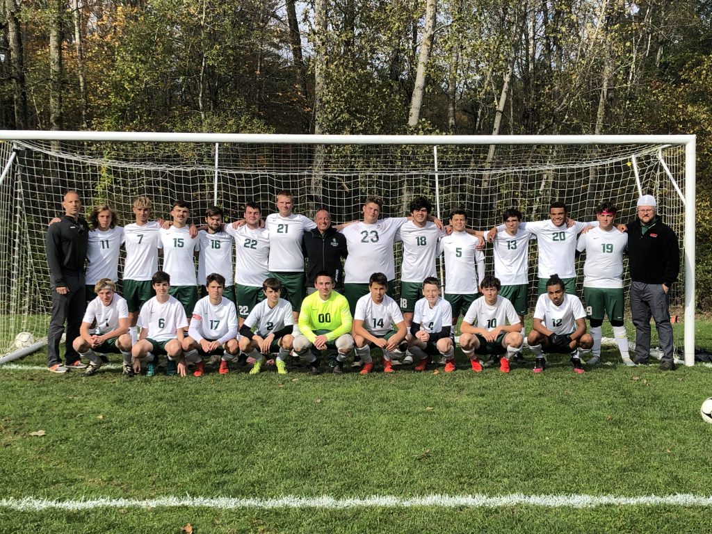 team picture of soccer players in white shirts and green shorts, the goalie is wearing a yellow shirt and the three coaches are wearing black