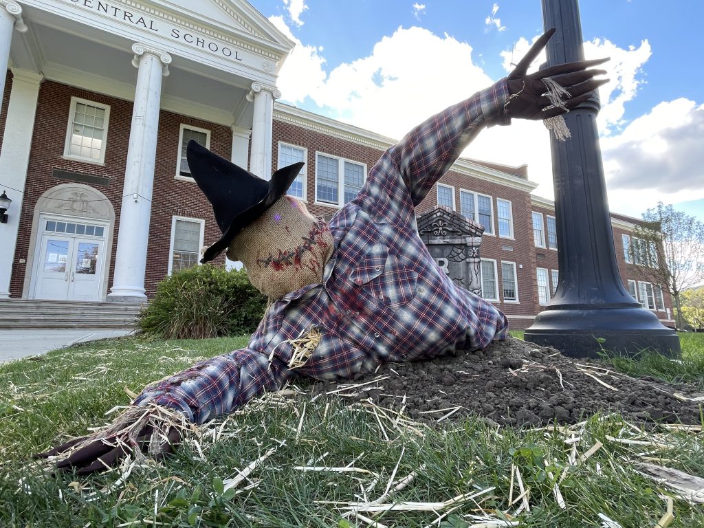 scarecrow with a flannel shirt and witches hat coming out of the ground and grave