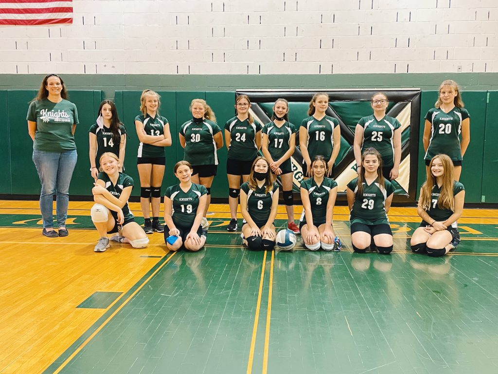 a coach stands next to nine volleyball players in uniform, six other players in uniform kneel in front of them