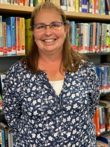 woman wearing glasses, a blue and white floral shirt stands in front of a book case with a big smile