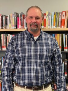 man with a goatee wearing a blue and white checkered shirt, standing in front of a book case