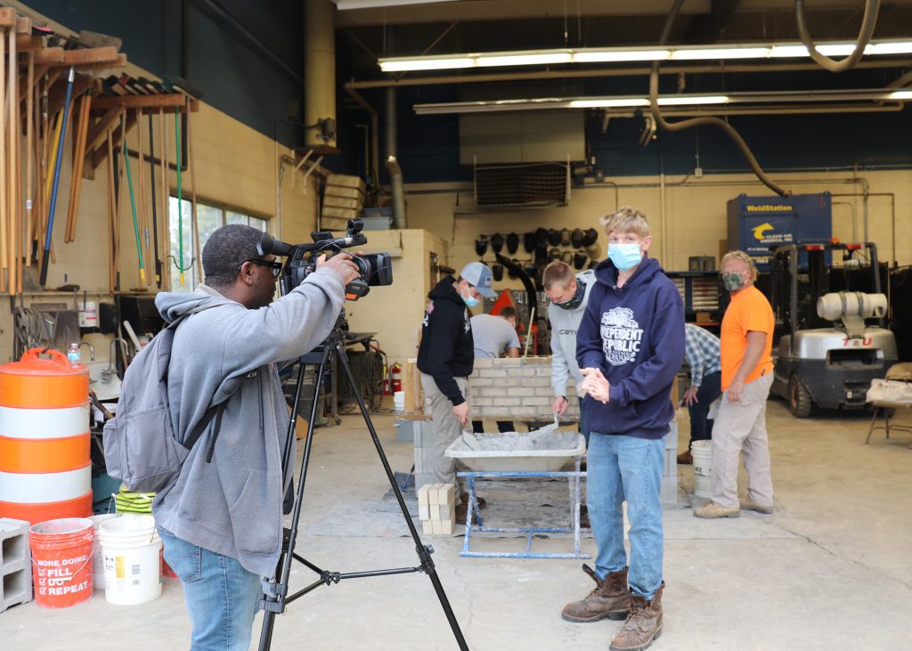 man behind camera and student in front of camera being interviewed. Other students are laying brick behind them