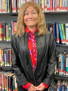 woman with blonde shoulder length hair smiling in front of a book case, wearing a black leather jacket and red floral shirt