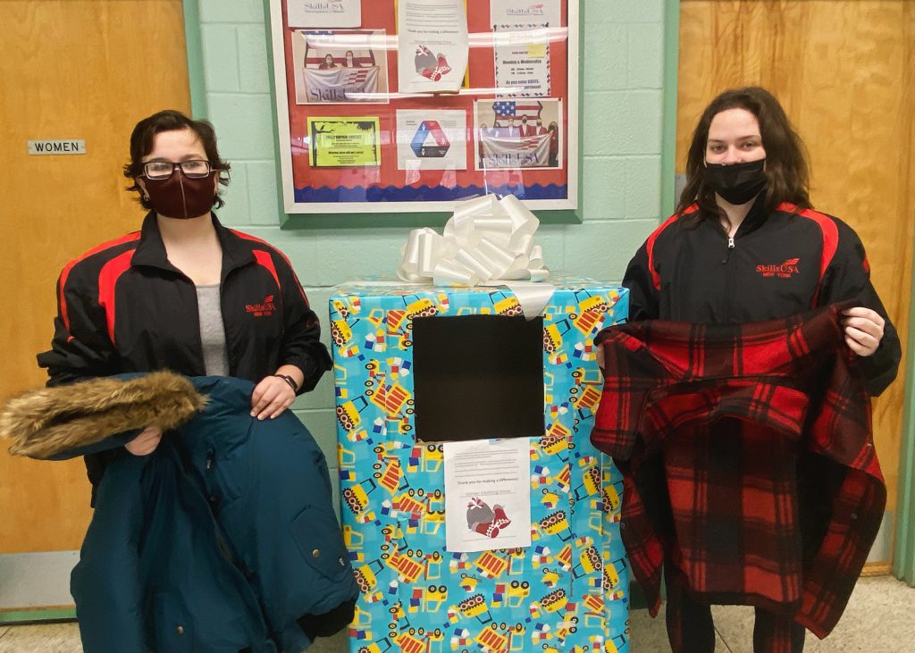 two students stand holding winter coats with a donation box in between them 