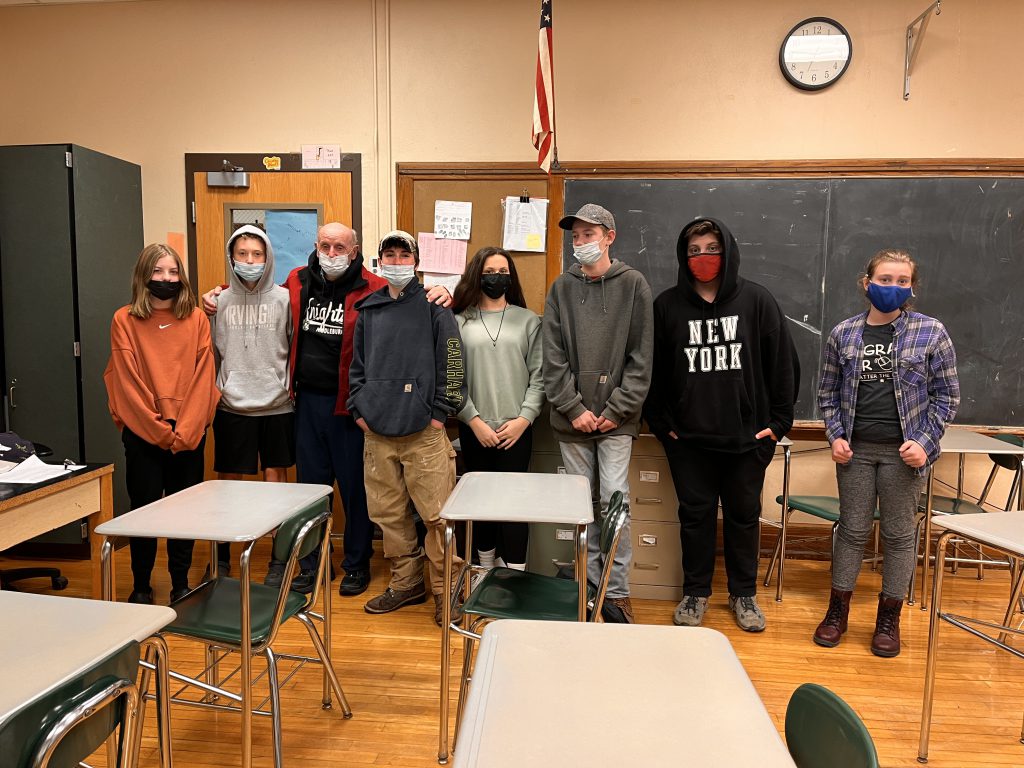 seven students stand around an army veteran in a classroom by the American flag