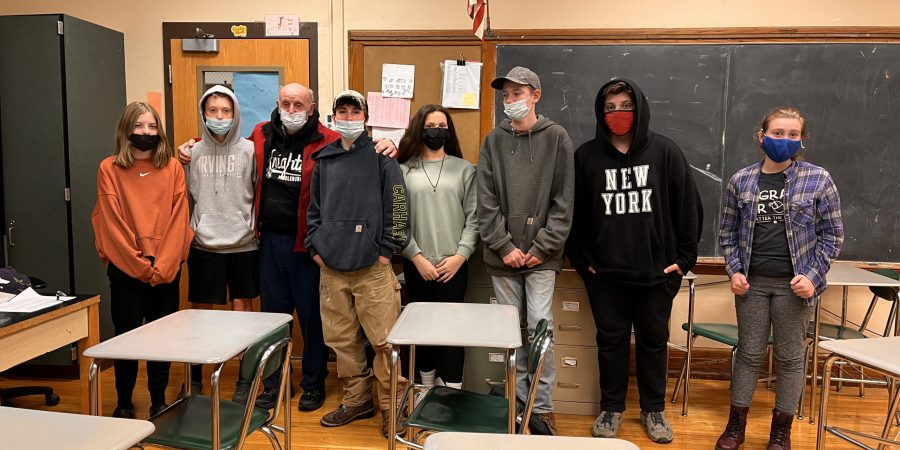 seven students stand around an army veteran in a classroom by the American flag