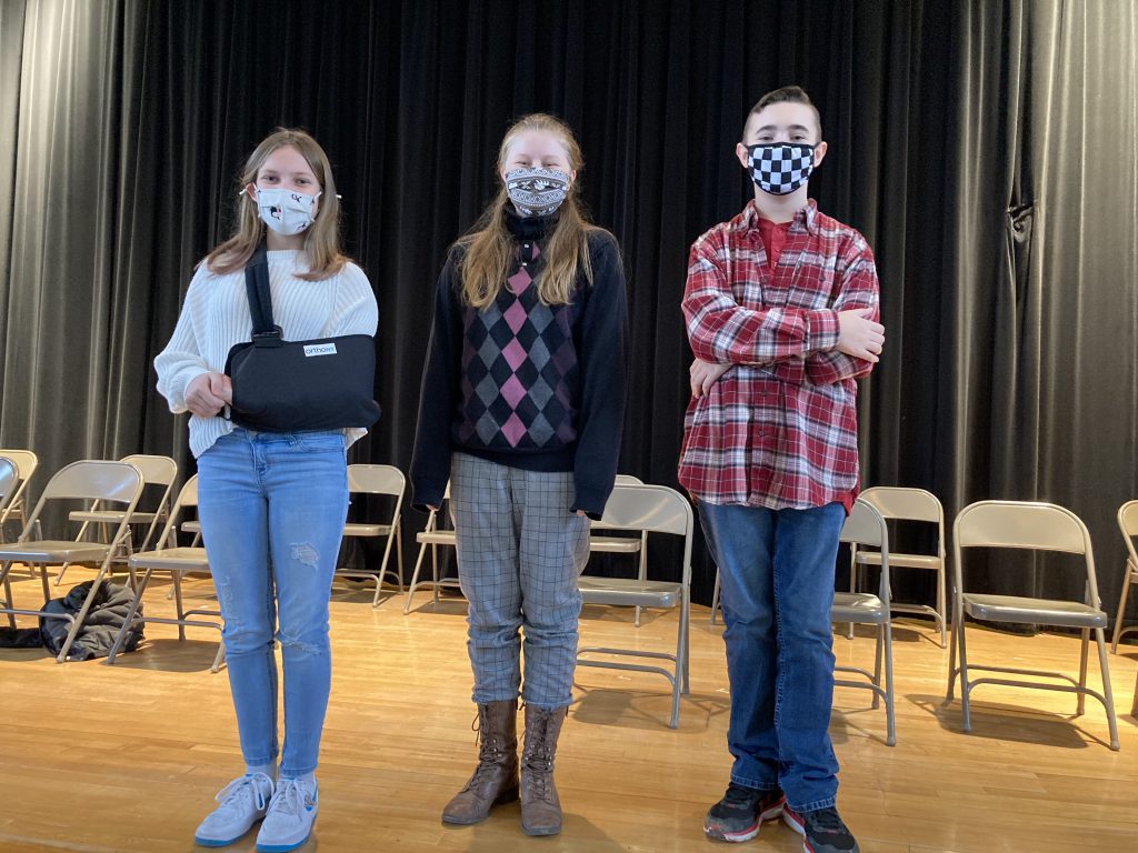 Three students standing on stage all wearing face masks. The first student has her arm in a sling. The second student is wearing an argyle sweater and the third student is wearing a flannel shirt