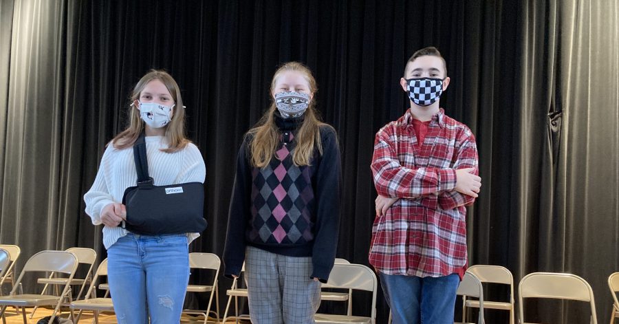 Three students standing on stage all wearing face masks. The first student has her arm in a sling. The second student is wearing an argyle sweater and the third student is wearing a flannel shirt.