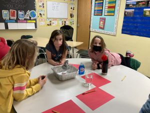 three students leaning on a table with red sheets of paper and a silver bowl in the middle