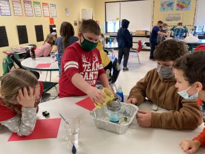 a classroom doing group projects and close to the screen are four students looking over a volcano experiment