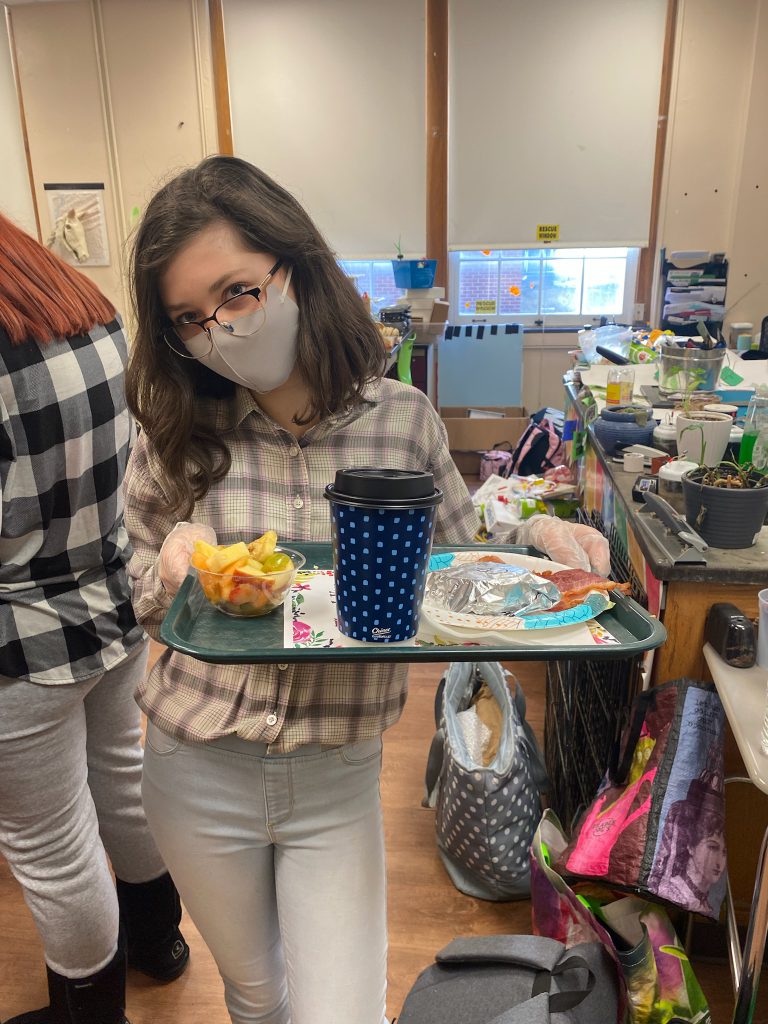 student holding a tray with fruit salad, bacon, a breakfast sandwich, and coffee