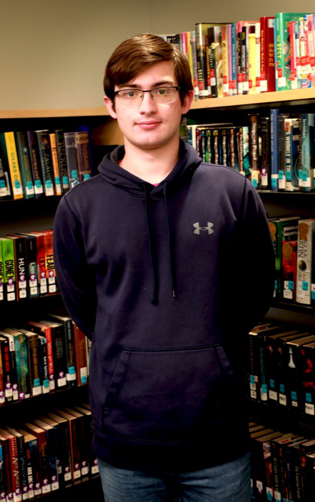 student with glasses and a navy hoodie smiles in front of bookcase