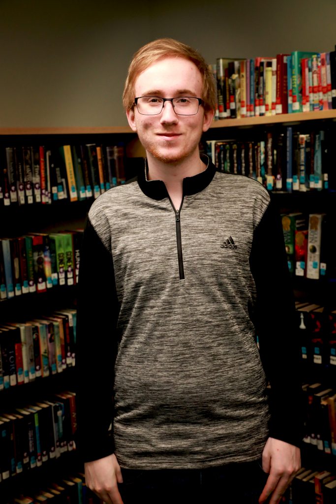 student with light hair and glasses stands proud in the library