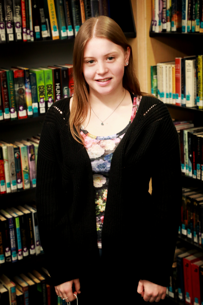 student with long, straight, red hair smiles in front of a bookcase