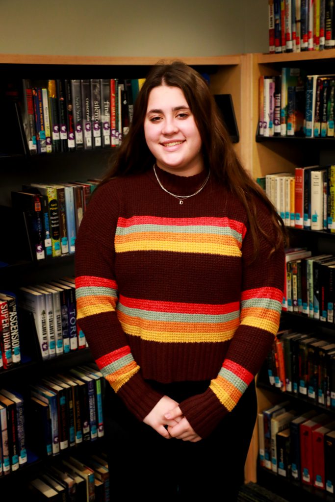 student with long brown hair wearing a brown, orange, grey and yellow striped sweater standing in front of a bookcase