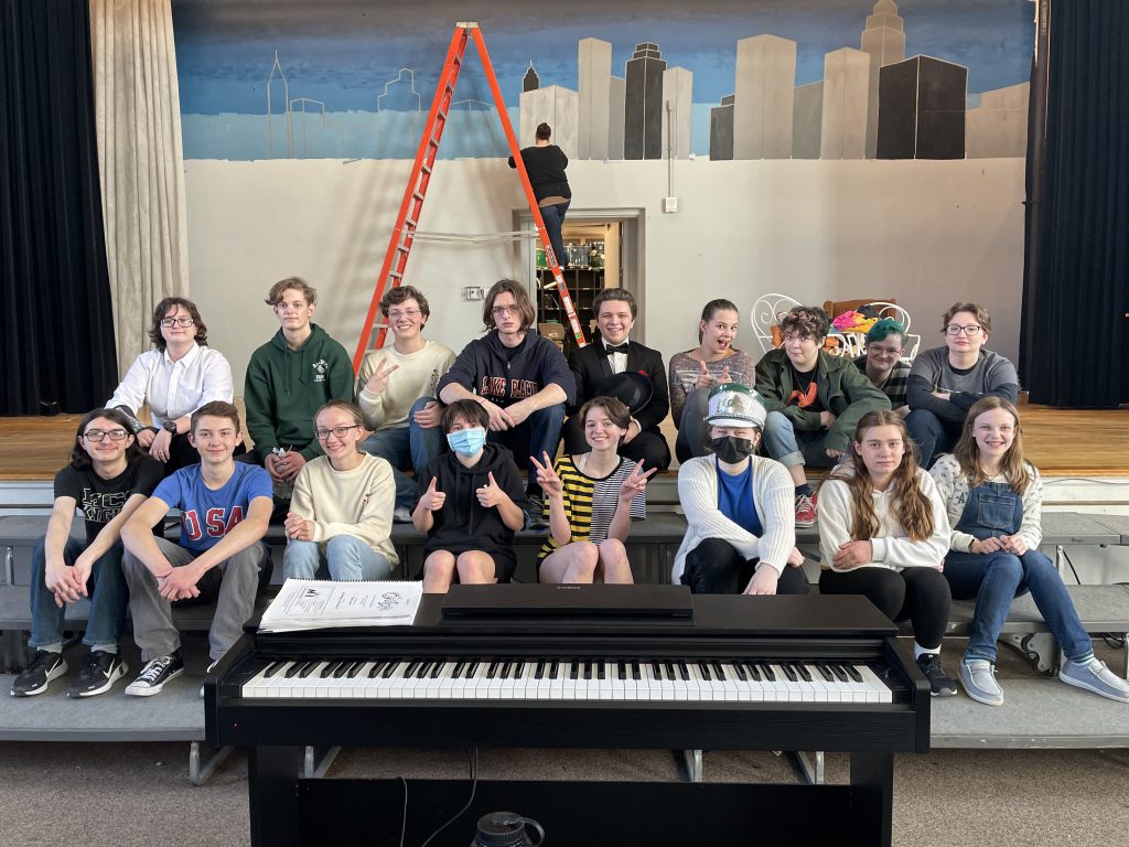 17 cast members smile on stage while a set designer paints in the background