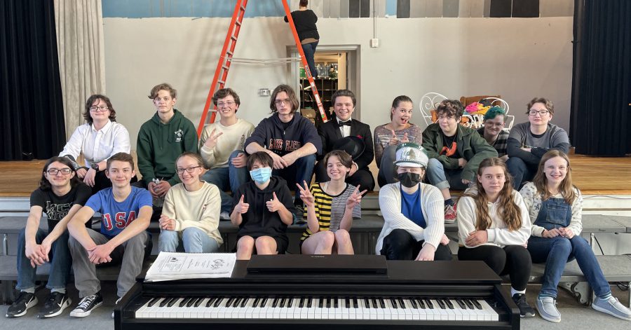 17 cast members smile on stage while a set designer paints in the background