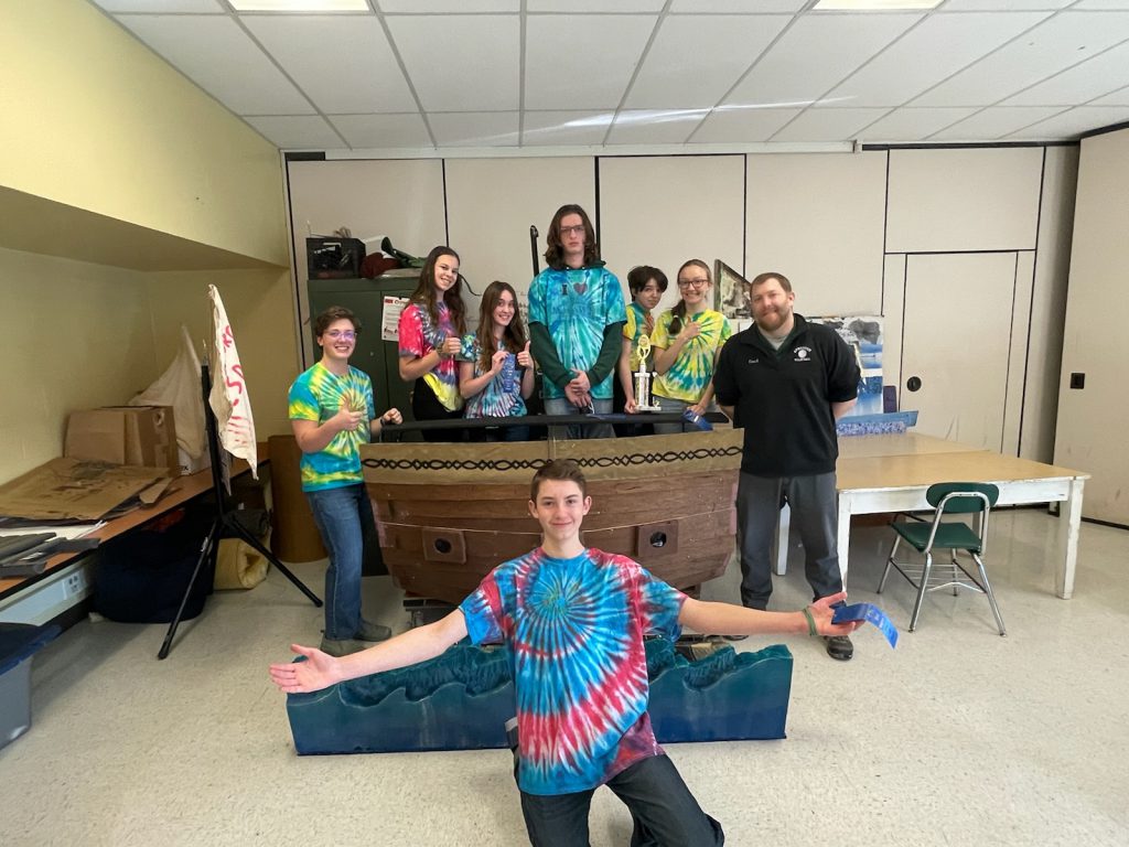 seven people behind a desk with one person kneeling in front with his arms out wide