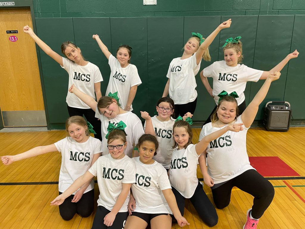 11 students wearing white shirts with MCS on it pose with arms out cheerleader-style