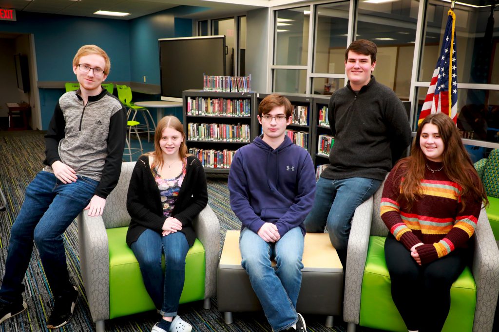 three students are sitting and two are leaning on the arms of the chairs in the library