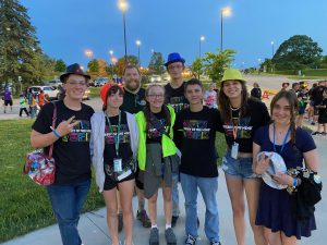 six students standing arm in arm with two others behind them in a parking lot with light posts on