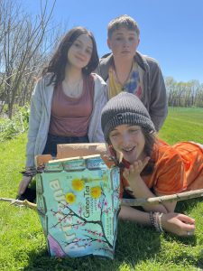 two students kneeling on the grass and one student laying down in front of an iced tea box