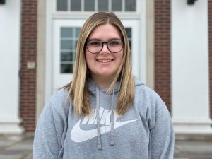 student wearing a grey Nike shirt, glasses, and blonde hair smiles in front of a brick school with white pillars