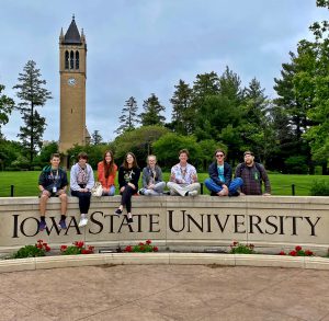 seven students sitting on a sign for Iowa State University and their coach is standing behind the sign