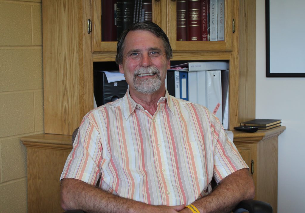 man with an orange, yellow, and white shirt sits with arms on chair and smiles