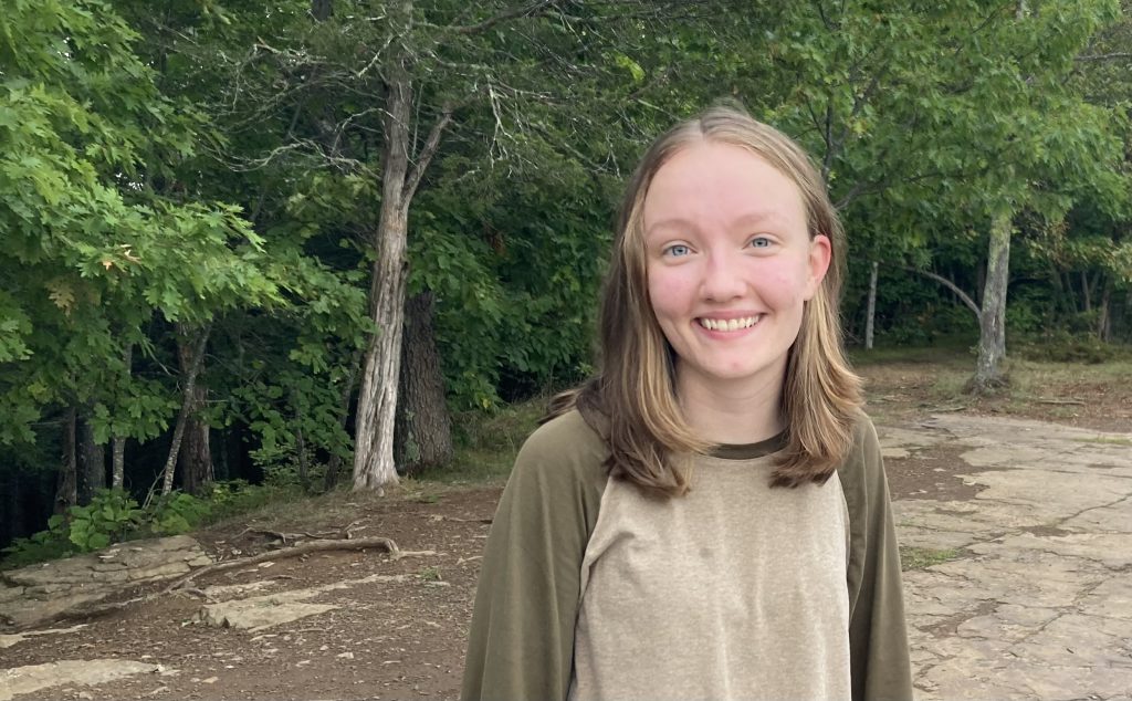 Sienna smiles at the camera. Her brown hair hangs down to her shoulders. Trees are in the background